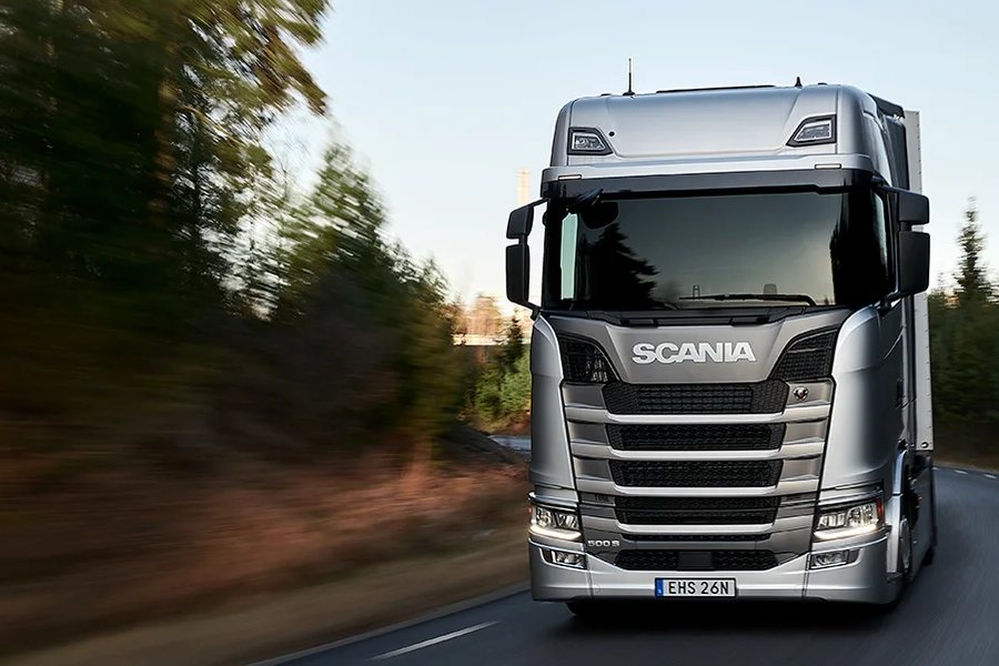 Camión Scania plateado circulando por una ruta entre árboles